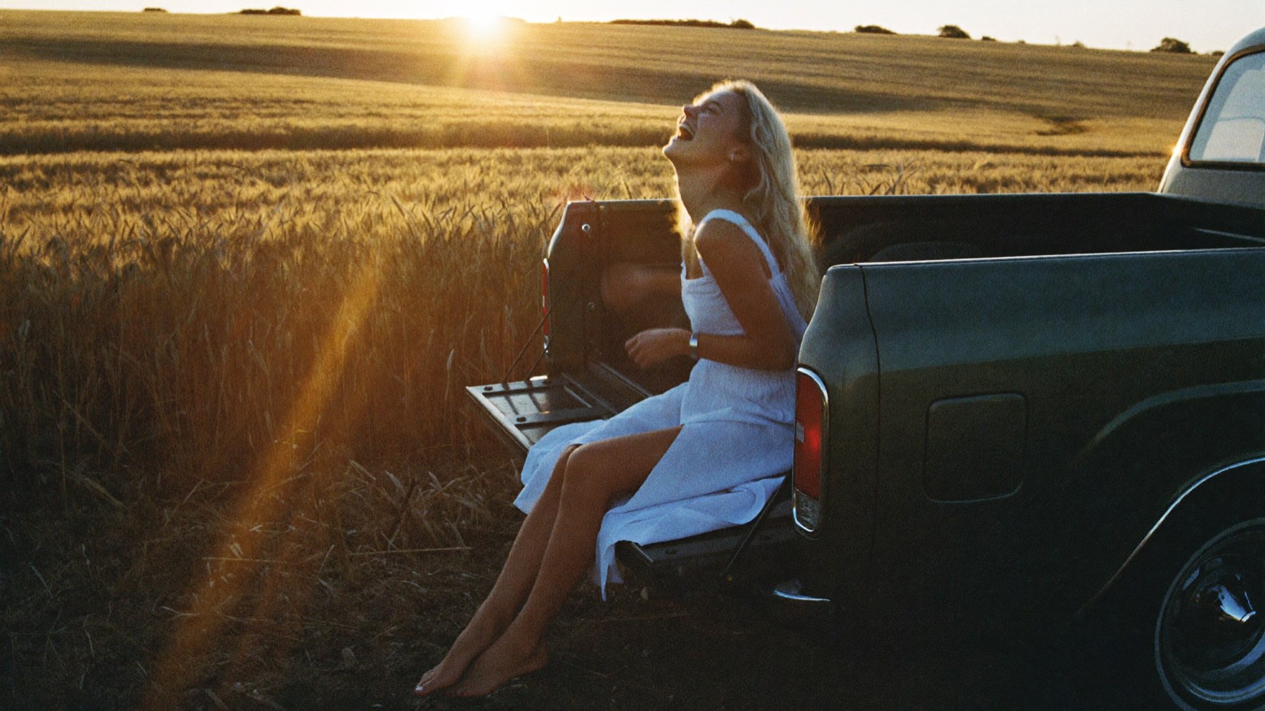 A young blonde woman in a white sundress laughing on the tailgate of a vintage pickup truck beside a golden field at sunset